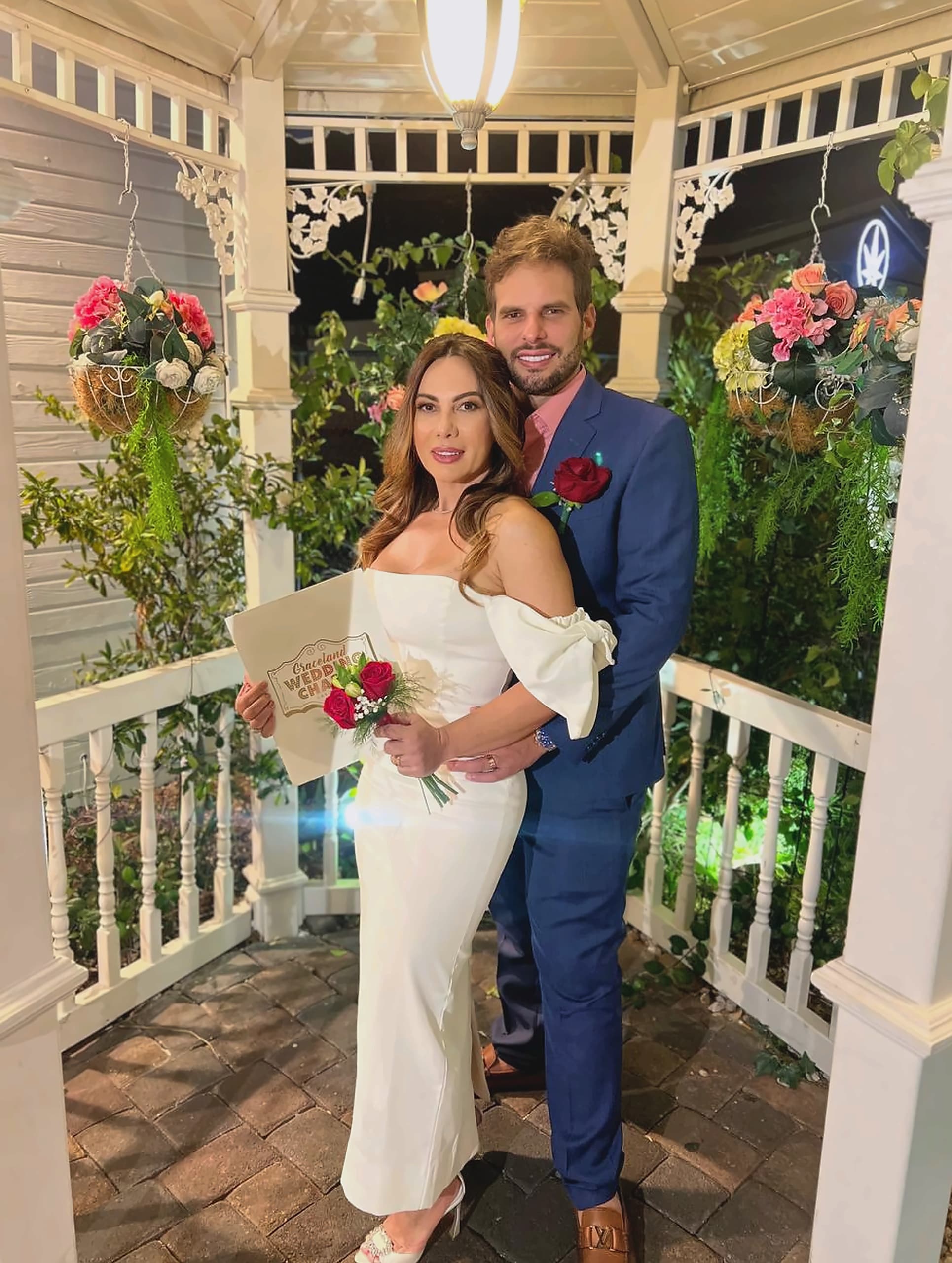 Gina e José Carlos, em traje de noiva e terno azul, abraçados em um gazebo decorado com flores.